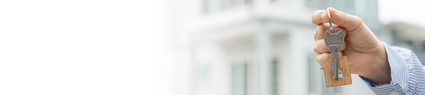 Hand holding keys with a house-shaped keychain in front of a white building