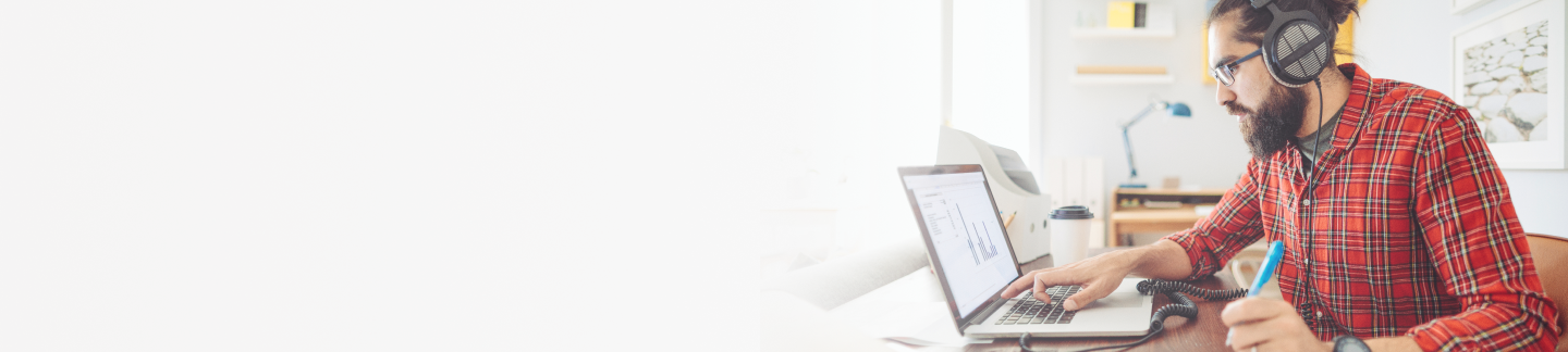 Man with a beard and glasses wearing headphones and a red plaid button down shirt looking at a laptop screen while writing notes with a pen in his left hand