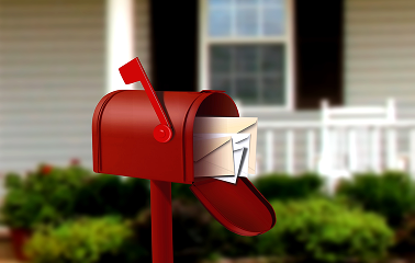 Red mailbox with flag up and several envelopes inside in front of a beige house with green shrubs and a white deck